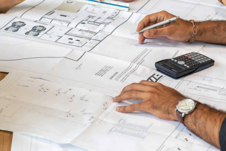 Mains avec calculatrice et crayon sur des plans étalés sur un bureau